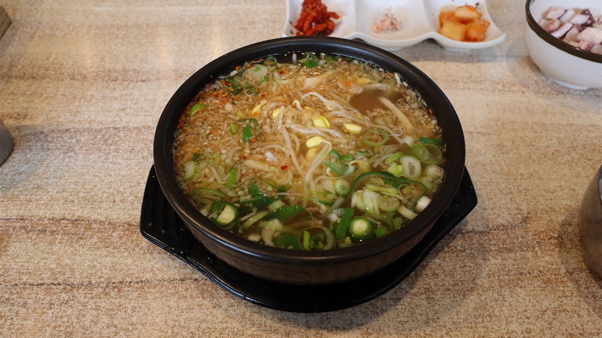 A black earthenware bowl of bean sprout soup with rice, topped with green onion, with side dishes